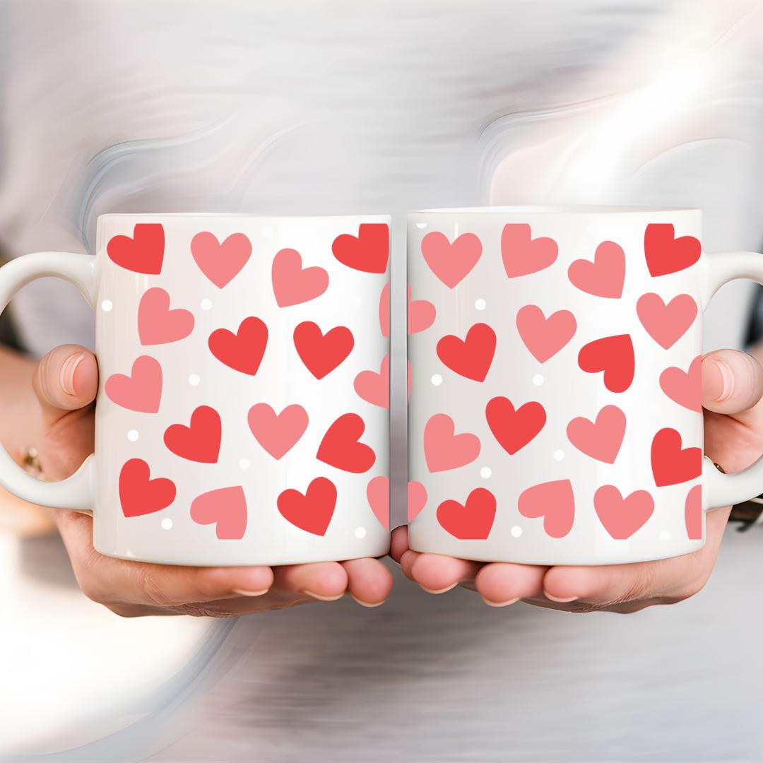 Weiße Keramiktasse mit roten und rosa Herzmustern, ergänzt durch weiße Punkte, gehalten von zwei Händen. Perfekt für Valentinstags- oder Alltagsmomente.